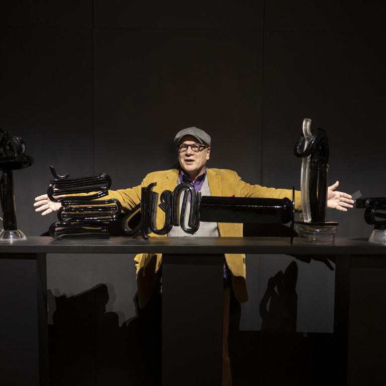 Izstādes telpā vīrietis cepurē un sinepju krāsas žaketē stāv aiz galda ar melnām stikla skulptūrām, izplētis rokas kā performatīvā pozā.