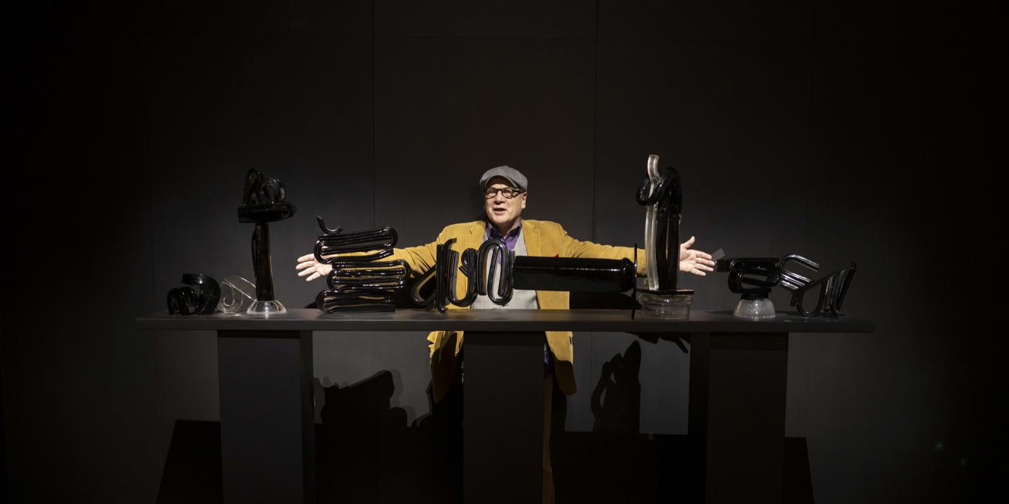 Izstādes telpā vīrietis cepurē un sinepju krāsas žaketē stāv aiz galda ar melnām stikla skulptūrām, izplētis rokas kā performatīvā pozā.