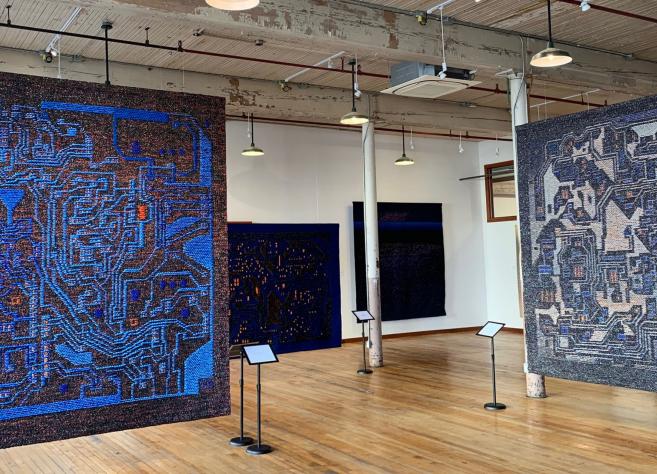 Izstāžu zālē redzami vairāki liela formāta tekstilmākslas darbi zilā un tumšos toņos, kas brīvi karājas telpā uz koka grīdas industriālā interjerā.