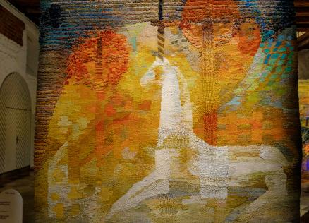 Izstādes telpā redzams liela mēroga tekstilmākslas darbs siltos dzeltenos, oranžos un zaļos toņos ar stilizētu balta zirga figūru kompozīcijas centrā.