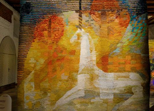 Izstādes telpā redzams liela mēroga tekstilmākslas darbs siltos dzeltenos, oranžos un zaļos toņos ar stilizētu balta zirga figūru kompozīcijas centrā.