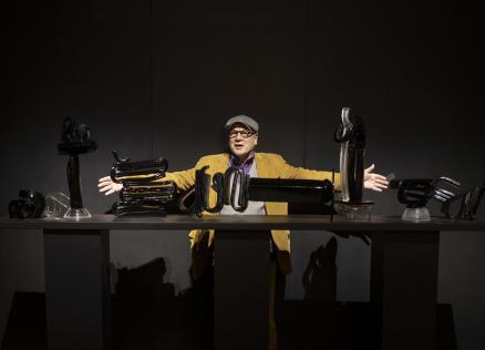 Izstādes telpā redzams liela mēroga tekstilmākslas darbs siltos dzeltenos, oranžos un zaļos toņos ar stilizētu balta zirga figūru kompozīcijas centrā.