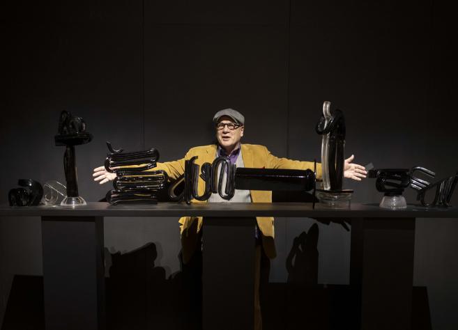 Izstādes telpā redzams liela mēroga tekstilmākslas darbs siltos dzeltenos, oranžos un zaļos toņos ar stilizētu balta zirga figūru kompozīcijas centrā.