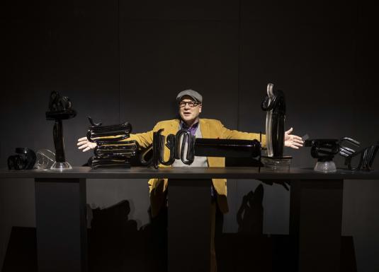 Izstādes telpā redzams liela mēroga tekstilmākslas darbs siltos dzeltenos, oranžos un zaļos toņos ar stilizētu balta zirga figūru kompozīcijas centrā.