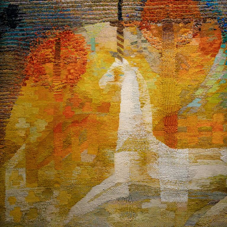 Izstādes telpā redzams liela mēroga tekstilmākslas darbs siltos dzeltenos, oranžos un zaļos toņos ar stilizētu balta zirga figūru kompozīcijas centrā.