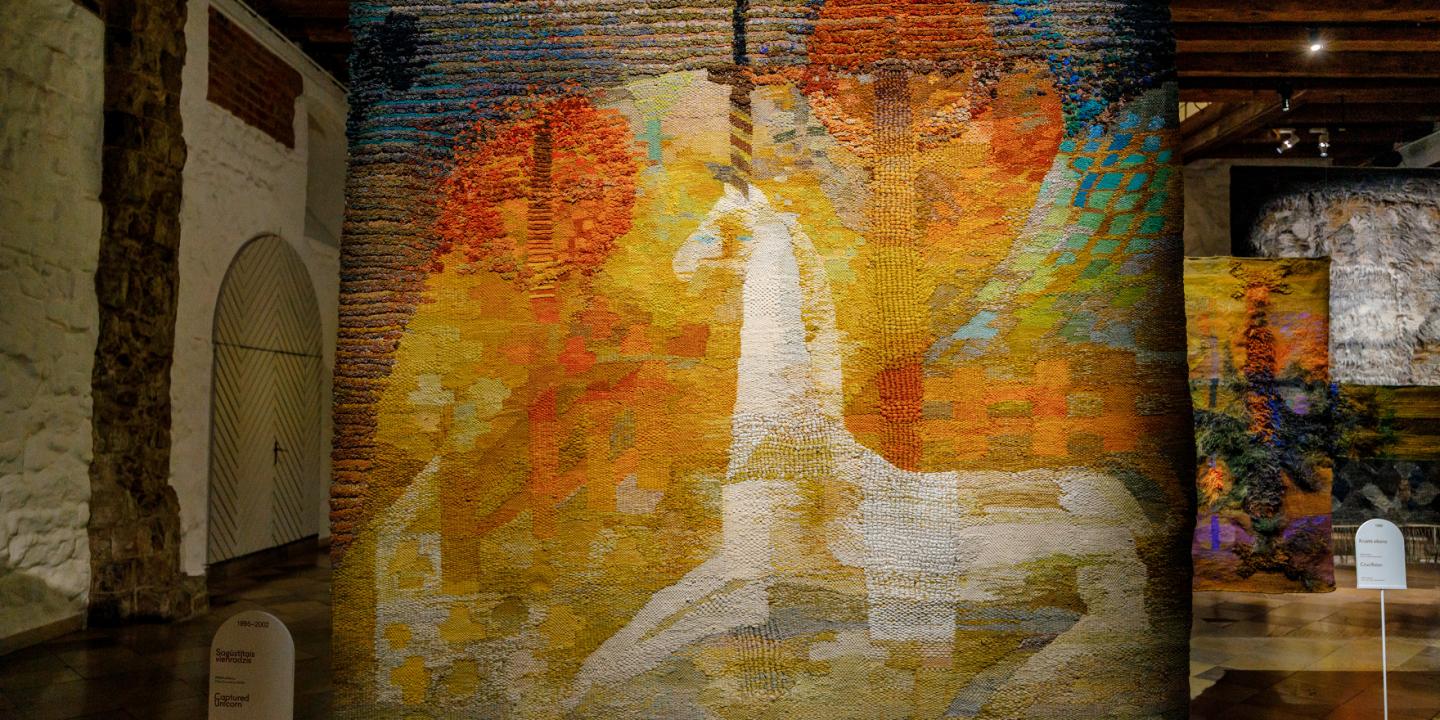 Izstādes telpā redzams liela mēroga tekstilmākslas darbs siltos dzeltenos, oranžos un zaļos toņos ar stilizētu balta zirga figūru kompozīcijas centrā.