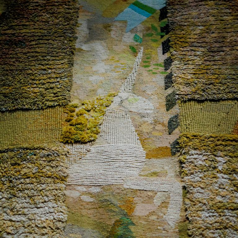 Tuvplāna skats uz tekstilmākslas darbu ar reljefu auduma virsmu un krustveida kompozīciju zemes toņos, izceļot aušanas faktūru un krāsu nianses.