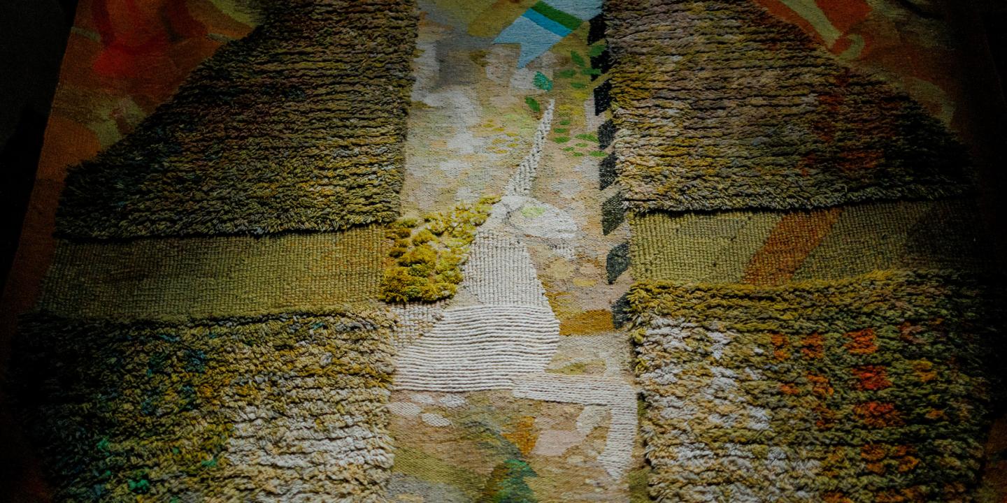 Tuvplāna skats uz tekstilmākslas darbu ar reljefu auduma virsmu un krustveida kompozīciju zemes toņos, izceļot aušanas faktūru un krāsu nianses.