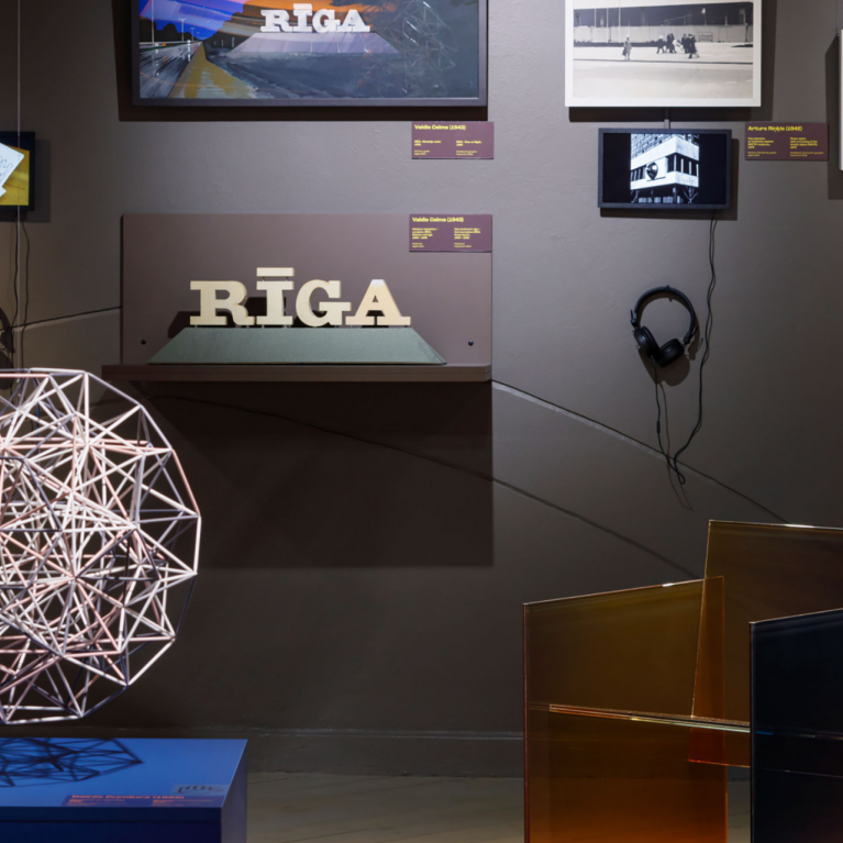 Izstādes ekspozīcijā redzams dizaina objektu, fotogrāfiju un multimediju darbu kopums ar izgaismotu telpisku struktūru priekšplānā un uzrakstu “RĪGA” centrā.
