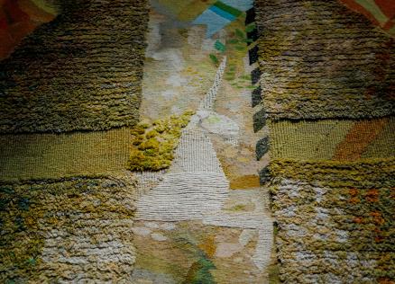 Tuvplāna skats uz tekstilmākslas darbu ar reljefu auduma virsmu un krustveida kompozīciju zemes toņos, izceļot aušanas faktūru un krāsu nianses.