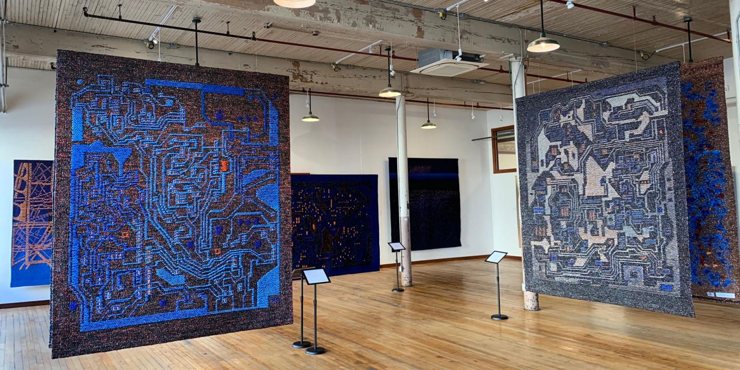 Izstāžu zālē redzami vairāki liela formāta tekstilmākslas darbi zilā un tumšos toņos, kas brīvi karājas telpā uz koka grīdas industriālā interjerā.