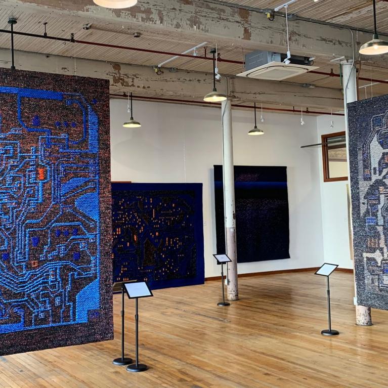 Izstāžu zālē redzami vairāki liela formāta tekstilmākslas darbi zilā un tumšos toņos, kas brīvi karājas telpā uz koka grīdas industriālā interjerā.