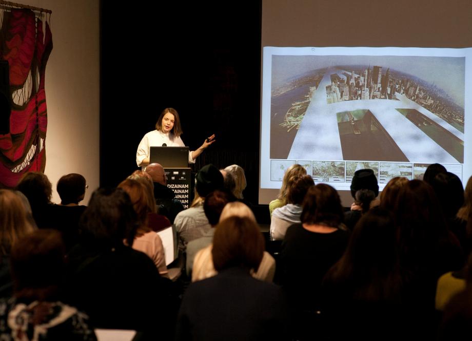 Lekcijas laikā runātāja auditorijai prezentē arhitektūras projektu ar pilsētas panorāmu, uz ekrāna redzams attēls, bet fonā pie sienas eksponēts liela izmēra tekstilmākslas darbs.