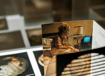 Attēlā redzama fotogrāfija ar sievieti, kas sēž gultā pie naktslampas un skatās portatīvajā datorā, izvietota izstādes ekspozīcijā uz galda ar citiem darbiem.