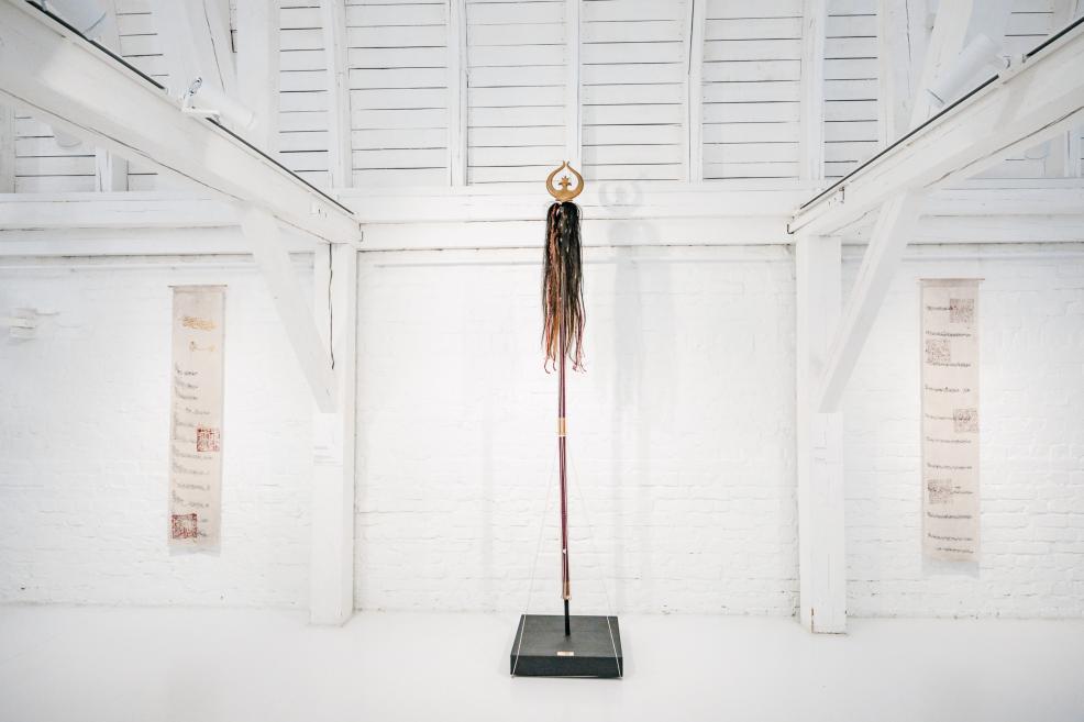 Installation view in a bright white gallery featuring a tall vertical sculpture with hanging strands at the center.