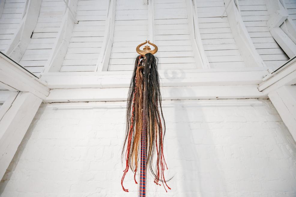 Close-up of a vertical sculpture topped with a golden crescent ornament and long braided strands hanging downward.