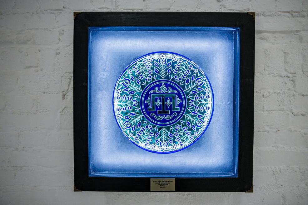 Illuminated circular ceramic plate with intricate blue and green patterns displayed on a white brick wall.