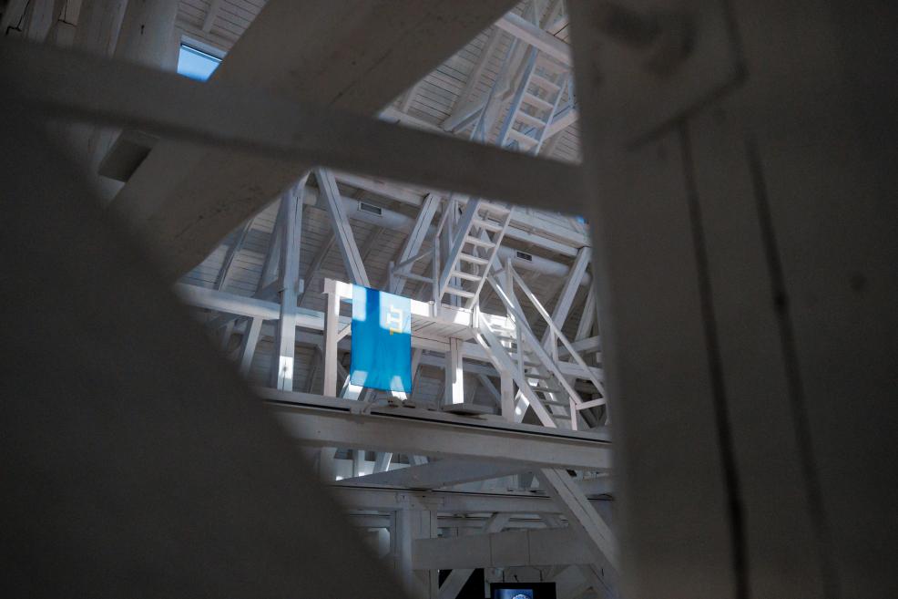 View through wooden beams revealing a hanging blue flag with a white symbol in a high-ceilinged white interior.