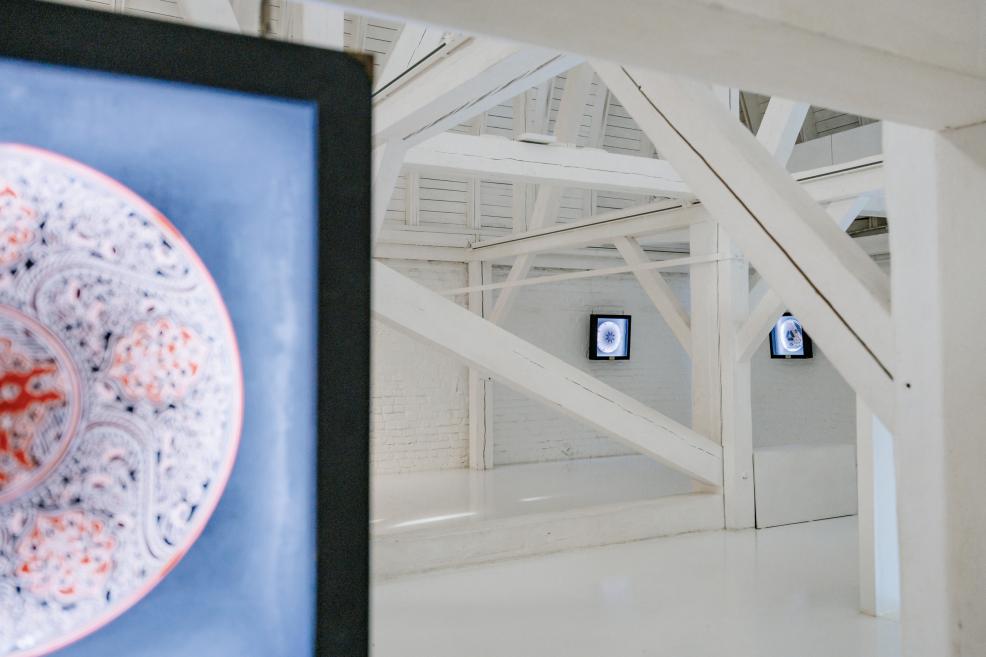 Exhibition view with illuminated ceramic plates on screens among white wooden beams.