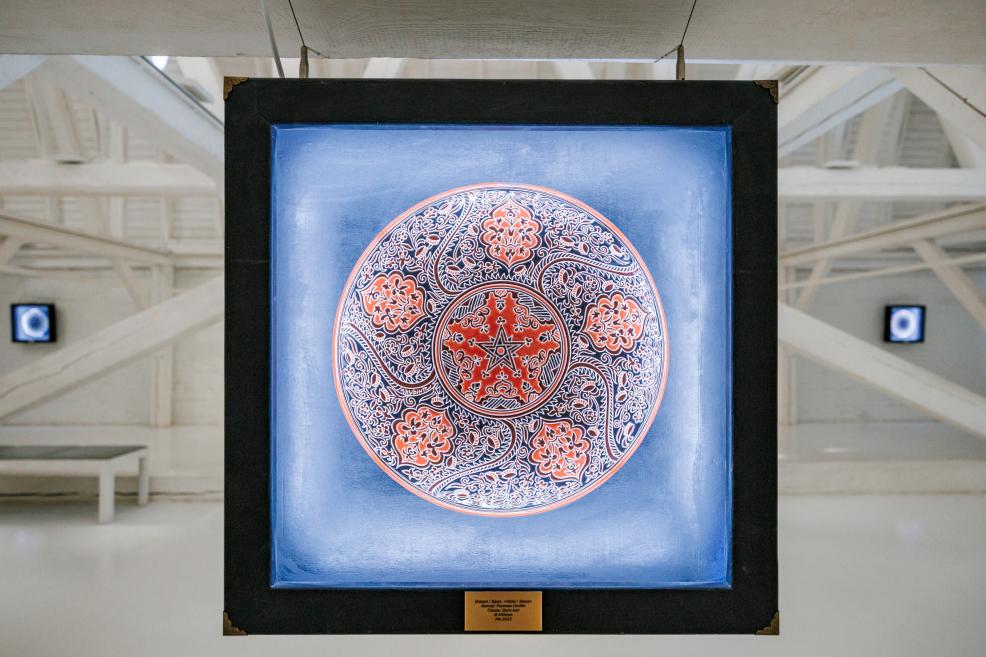 Illuminated red and blue patterned circular artwork displayed in a black frame against a white gallery background.