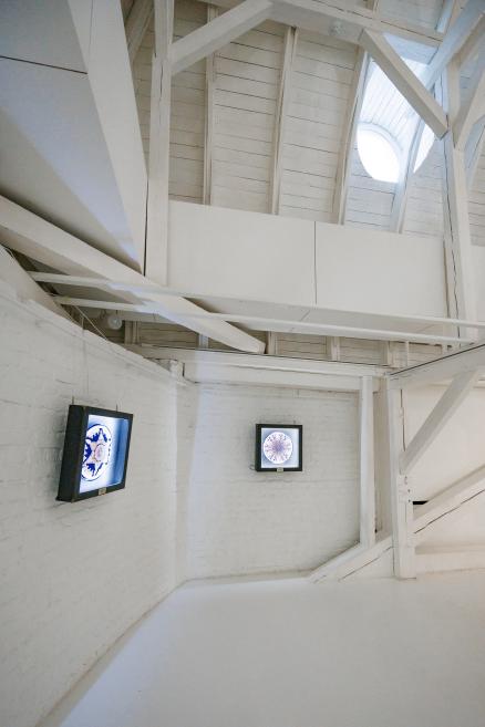 A white exhibition space with a tall pole sculpture topped with hair-like strands and symbols on the wall scrolls.
