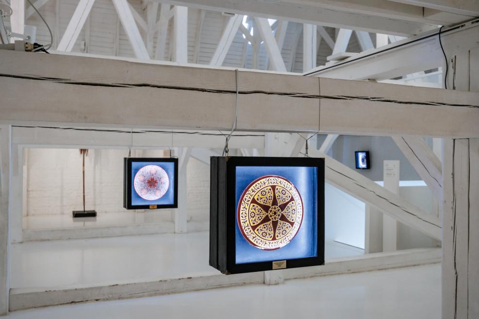 View of the exhibition hall with multiple illuminated ceramic plates suspended in a white space.