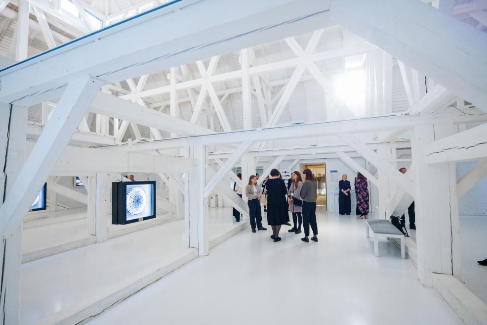 Cilvēki pulcējas un sarunājas gaišā galerijā ar piekārtiem apļveida mākslas darbiem.