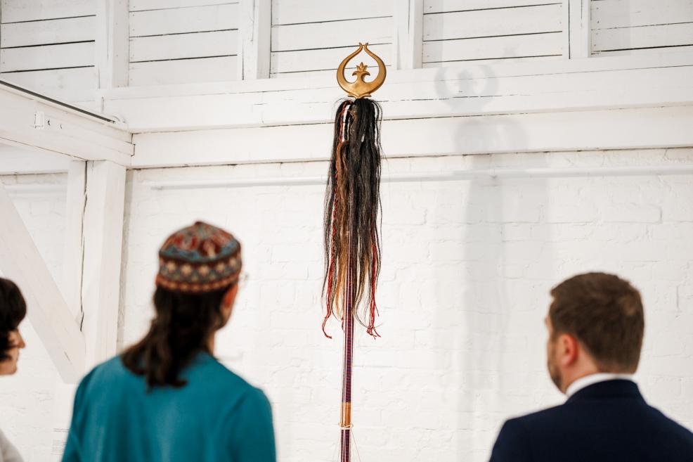 Izstādes apmeklētāji aplūko augstu skulpturālu objektu ar dekoratīvām lentēm un zelta augšdaļu.