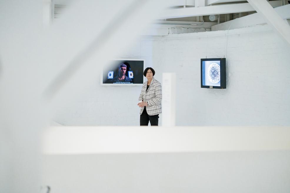 Sieviete stāv baltā izstāžu zālē, kur uz sienas redzami video un apļveida mākslas darbi.