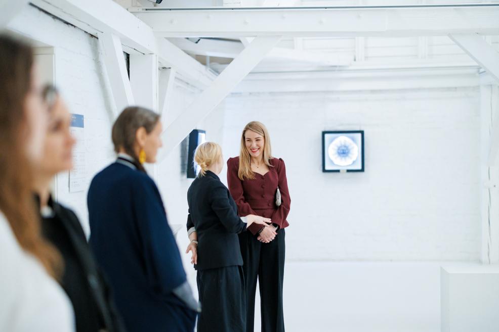 Apmeklētāji sarunājas gaišā baltā galerijā ar ģeometriskām griestu sijām un pie sienas izvietotiem mākslas darbiem.