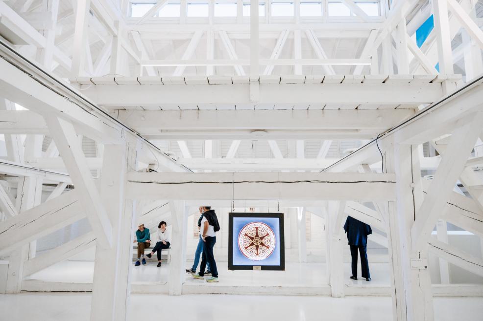 Cilvēki pulcējas gaišā, baltā galerijā ar karājošiem apļveida mākslas darbiem.