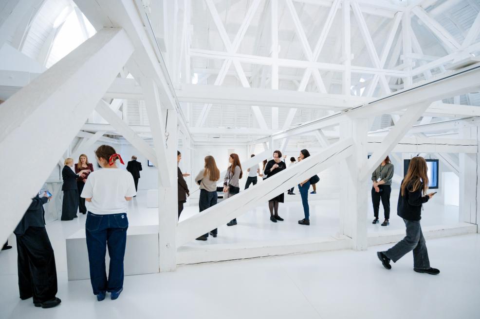 Apmeklētāji izpēta plašu baltu izstāžu zāli ar vairākiem mākslas darbiem un koka konstrukcijām.