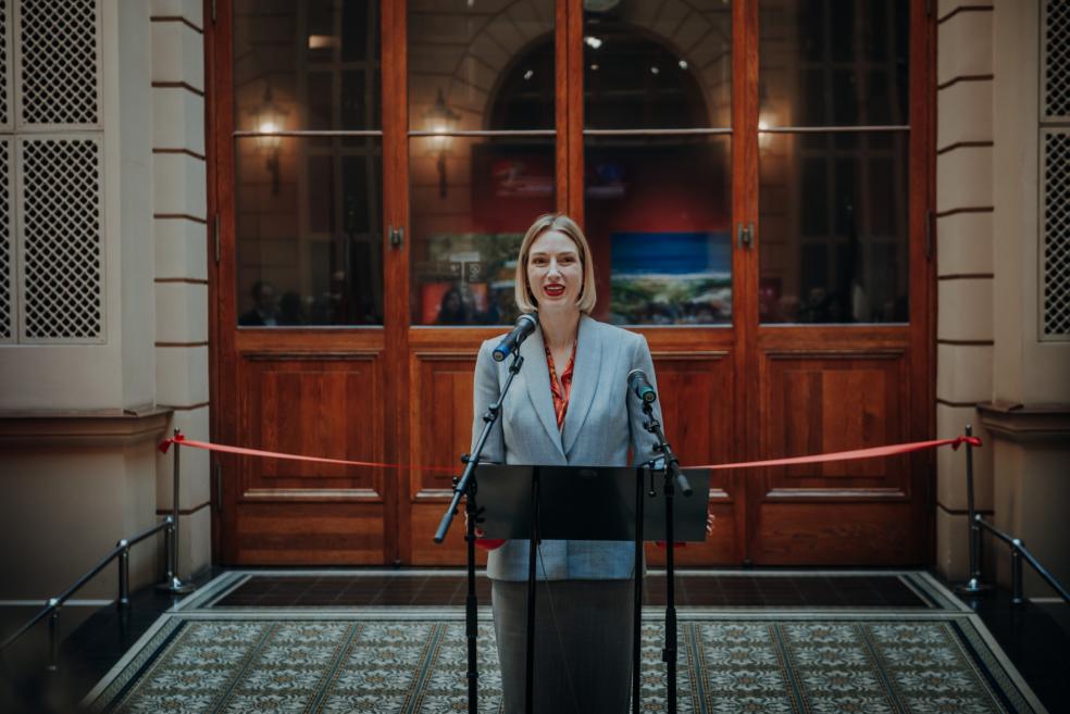 A woman in a grey suit with red accents speaks during the event.