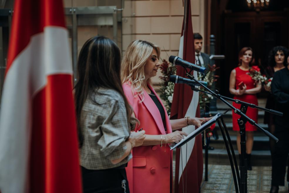 A woman in a pink coat speaks at the podium with microphones.