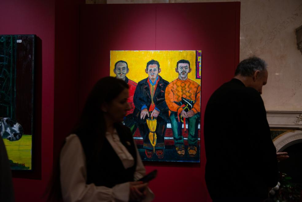 Visitors view an exhibition with vivid painting compositions displayed on red walls.