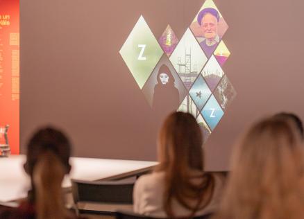 Apmeklētāji skatās prezentāciju ar vizuāliem attēliem uz ekrāna, kas veidoti dimanta formā, izstādē ar sarkanu sienu fonā un uzrakstu “Forma un materiāls”.