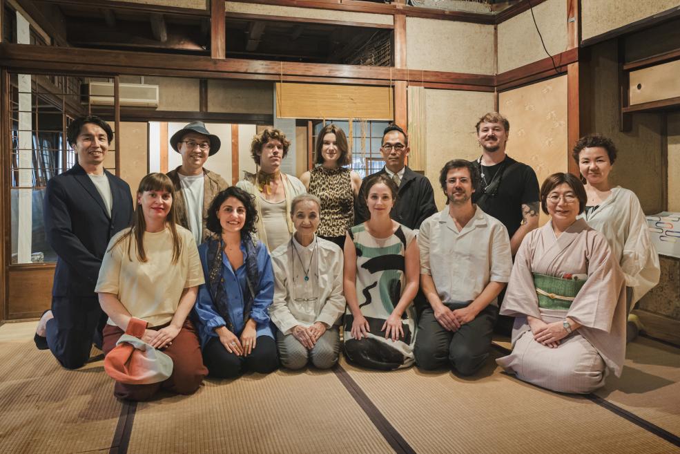 Cilvēku grupa pozē kopīgai fotogrāfijai tradicionālā japāņu interjerā ar tatami grīdu un koka sienām.