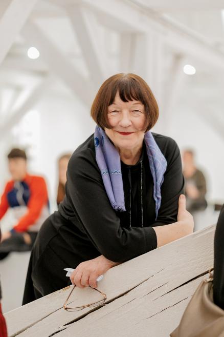 Sieviete melnā apģērbā ar violetu šalli atspiedusies pret margām, fonā izplūdusi galerijas telpa ar cilvēkiem.