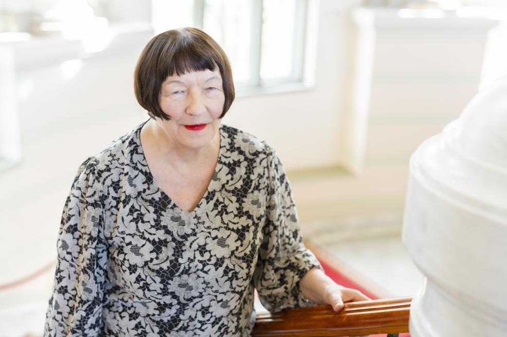 Sieviete rakstainā kleitā sēž gaišā galerijas telpā pie margām, fonā redzama skulptūra.