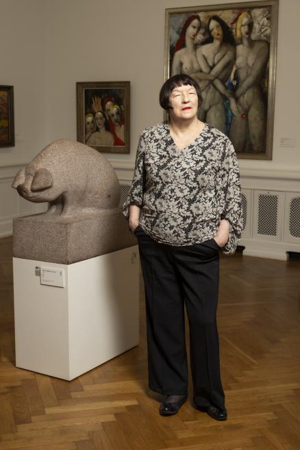 Sieviete stāv mākslas galerijā blakus akmens skulptūrai, fonā redzamas figurālas gleznas.