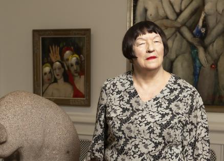 Sieviete stāv mākslas galerijā blakus akmens skulptūrai, fonā redzamas figurālas gleznas.