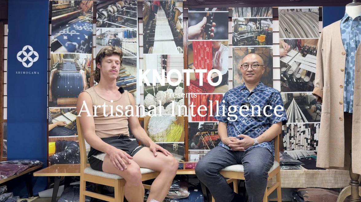 Divi vīrieši sēž uz krēsliem tekstila darbnīcā ar aušanas procesu fotogrāfijām fonā un uzrakstu “Artisanal Intelligence”.