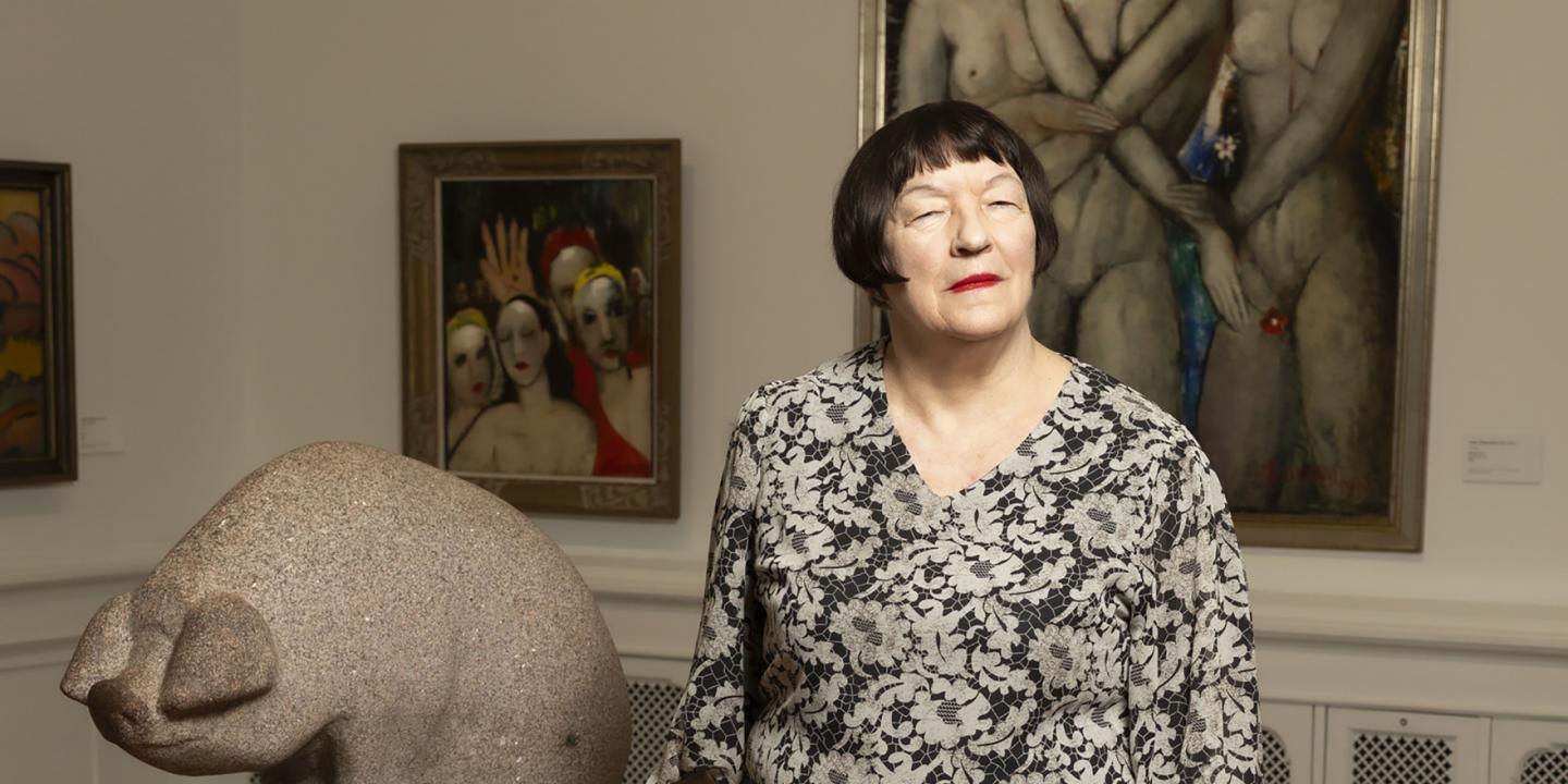 Sieviete stāv mākslas galerijā blakus akmens skulptūrai, fonā redzamas figurālas gleznas.