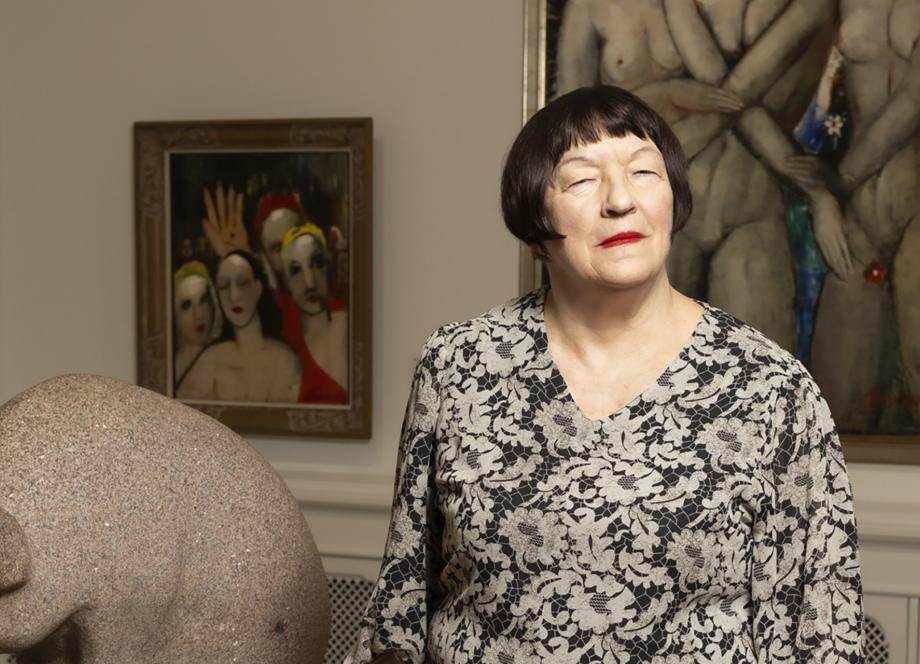 Sieviete stāv mākslas galerijā blakus akmens skulptūrai, fonā redzamas figurālas gleznas.