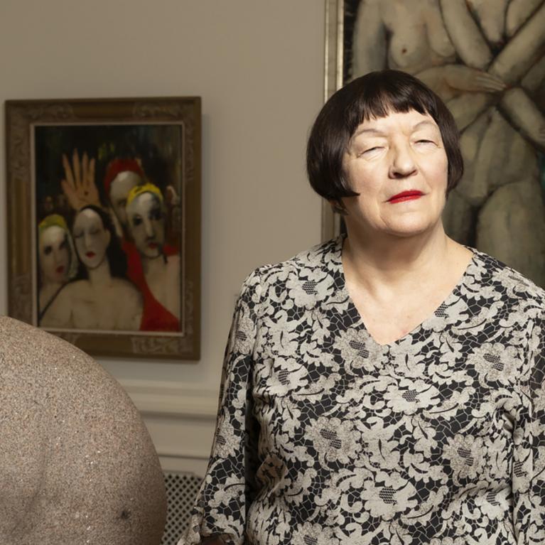 Sieviete stāv mākslas galerijā blakus akmens skulptūrai, fonā redzamas figurālas gleznas.