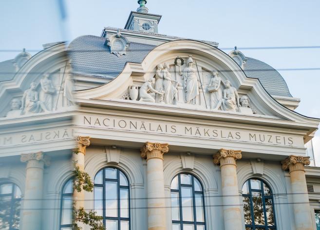 Latvijas Nacionālā mākslas muzeja vēsturiskā ēka ar monumentālu fasādi, skulpturālu frīzi un klasiskām kolonnām mirdz maigā dienas gaismā.