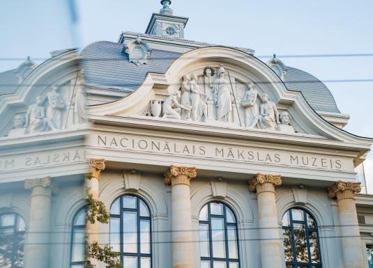 Latvijas Nacionālā mākslas muzeja vēsturiskā ēka ar monumentālu fasādi, skulpturālu frīzi un klasiskām kolonnām mirdz maigā dienas gaismā.