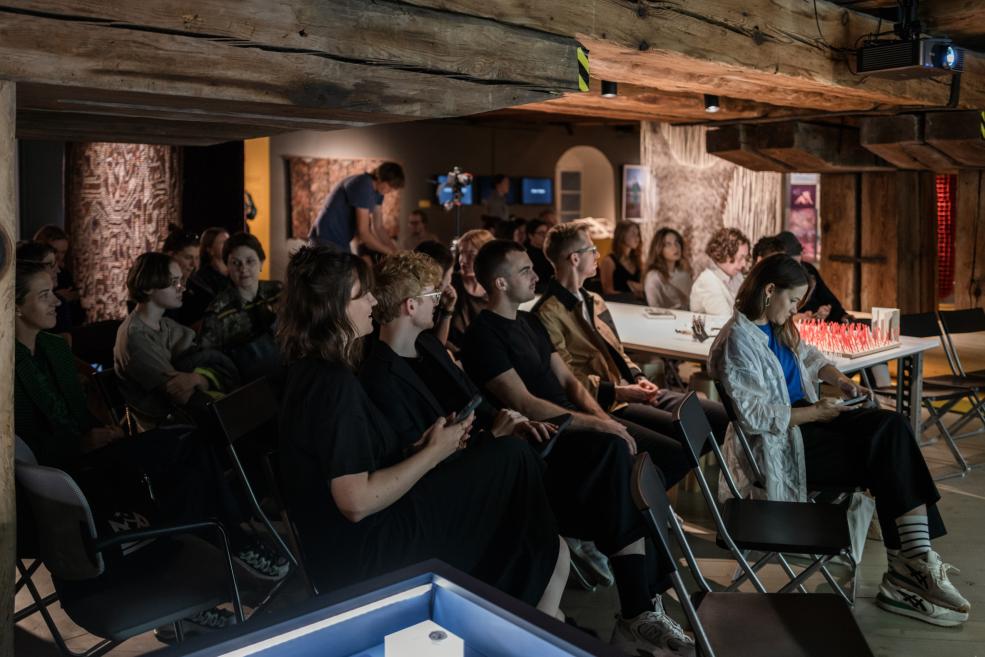 Muzeja zālē zem koka sijām sēž auditorija, kas klausās prezentāciju pie galdiem ar mākslas objektiem.