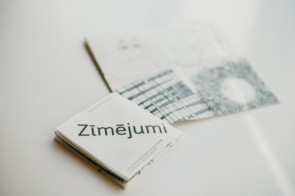 Uz balta fona redzama uz galda novietota atvērta brošūra ar uzrakstu Zīmējumi un blakus daļēji redzama vēl viena drukāta lapa ar zilu līniju režģi.