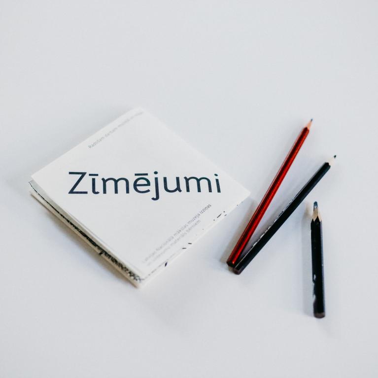 Balta grāmata ar uzrakstu Zīmējumi un trīs zīmuļi uz gaišax fona.